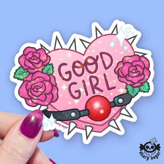 A hand with glittery purple nail polish holds a die-cut sticker featuring a pink heart with the words 'GOOD GIRL' in dark pink letters, pink roses with green leaves, and a red heart-shaped lock with a black key. The sticker is set against a light blue background with a 'sharp sugar' brand logo in the bottom right corner.