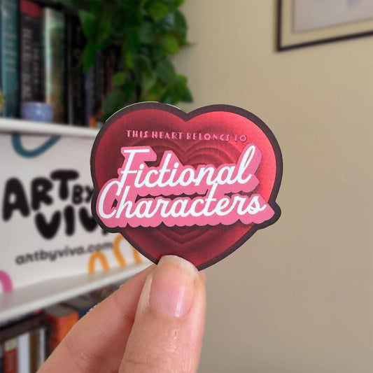 Heart-shaped sticker with text held by a hand in front of a bookshelf.