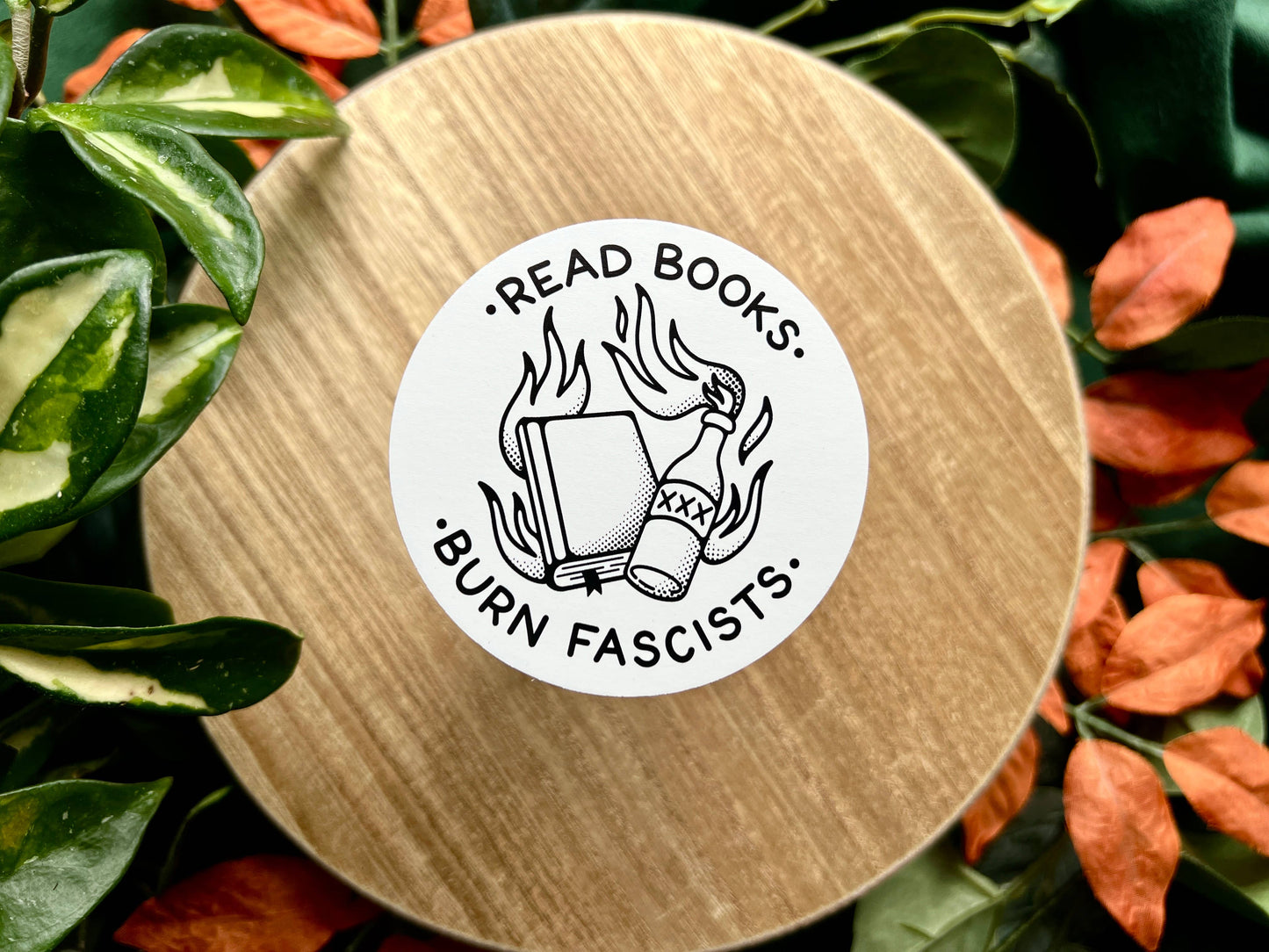 Round wooden coaster with a sticker reading 'Read books. Burn fascists.' surrounded by leaves.