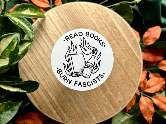 Round wooden coaster with a sticker reading 'Read books. Burn fascists.' surrounded by leaves.