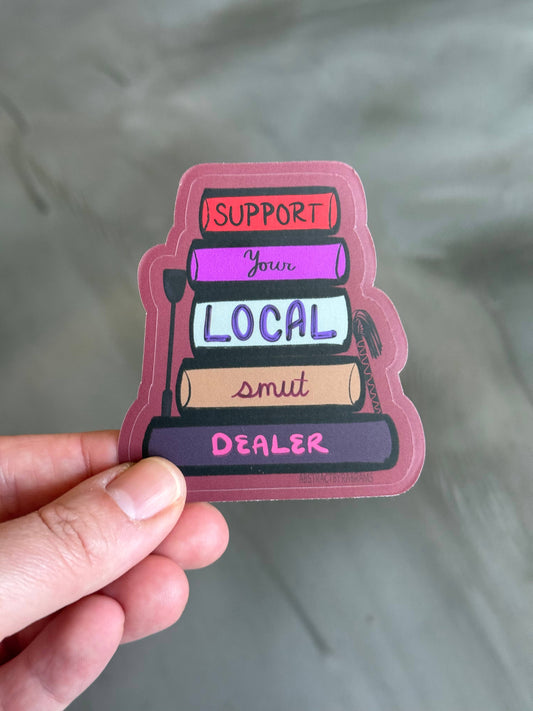 Sticker with text 'Support Your Local Smut Dealer' held by a hand against a gray background