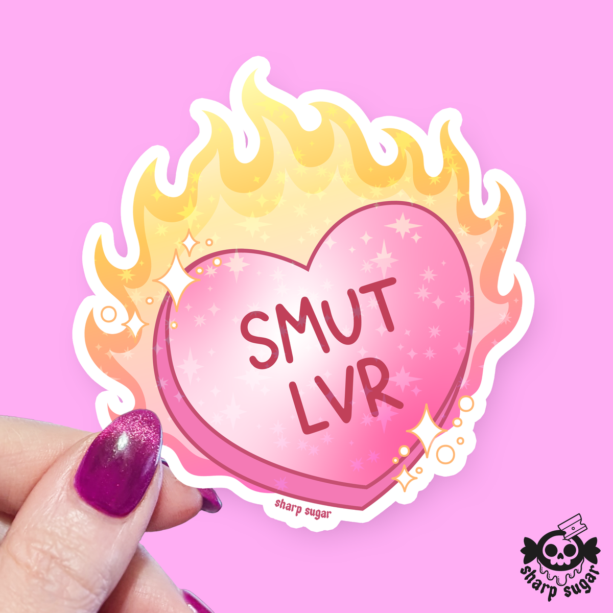 A die-cut sticker held by a hand with glittery purple nail polish. The sticker features a pink heart with the text 'SMUT LVR' in dark pink, surrounded by yellow and orange flames with small white stars.
