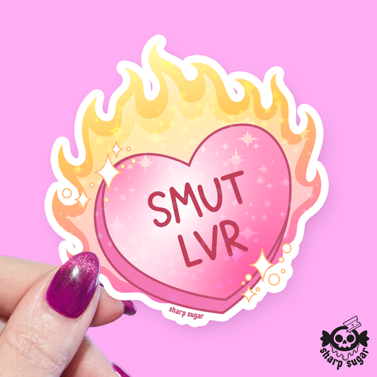 A die-cut sticker held by a hand with glittery purple nail polish. The sticker features a pink heart with the text 'SMUT LVR' in dark pink, surrounded by yellow and orange flames with small white stars.