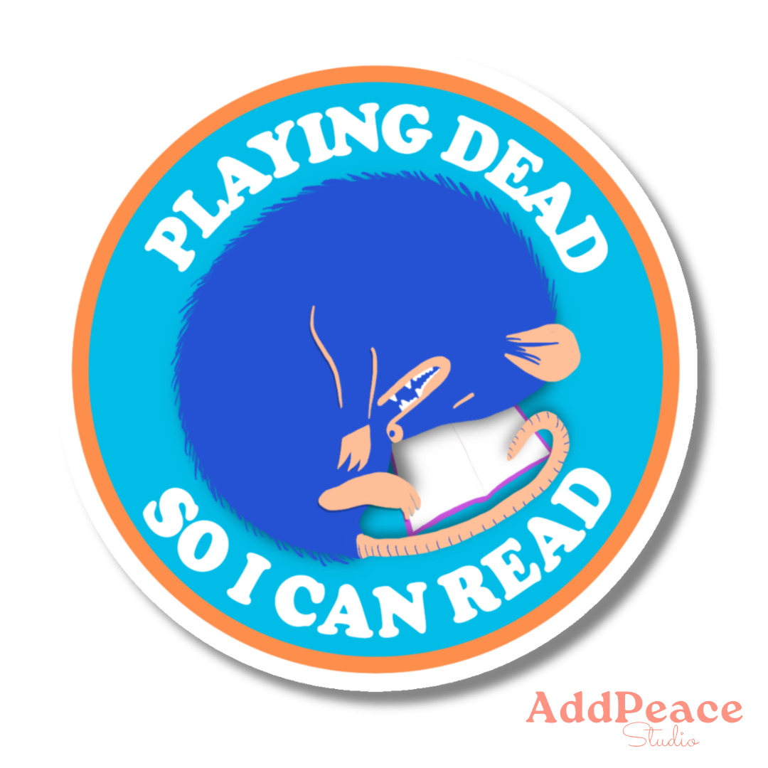 Circular sticker with blue character reading a book and text 'Playing dead so I can read' on a white background.