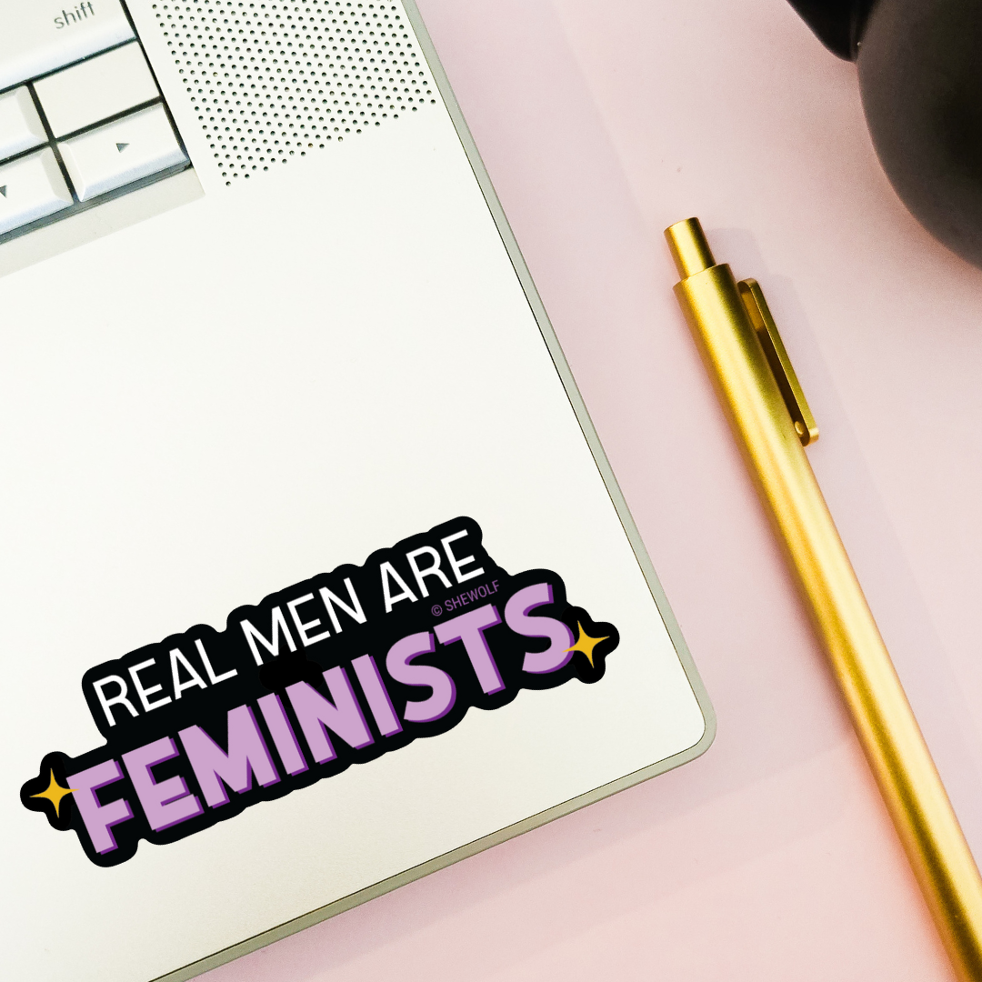 Notebook with 'Real Men Are Feminists' sticker on a pink surface with a gold pen.