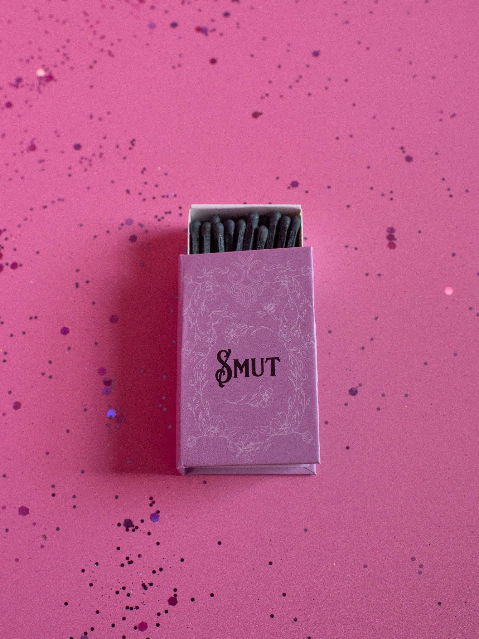 Matchbox labeled 'Smut' on a pink background with confetti