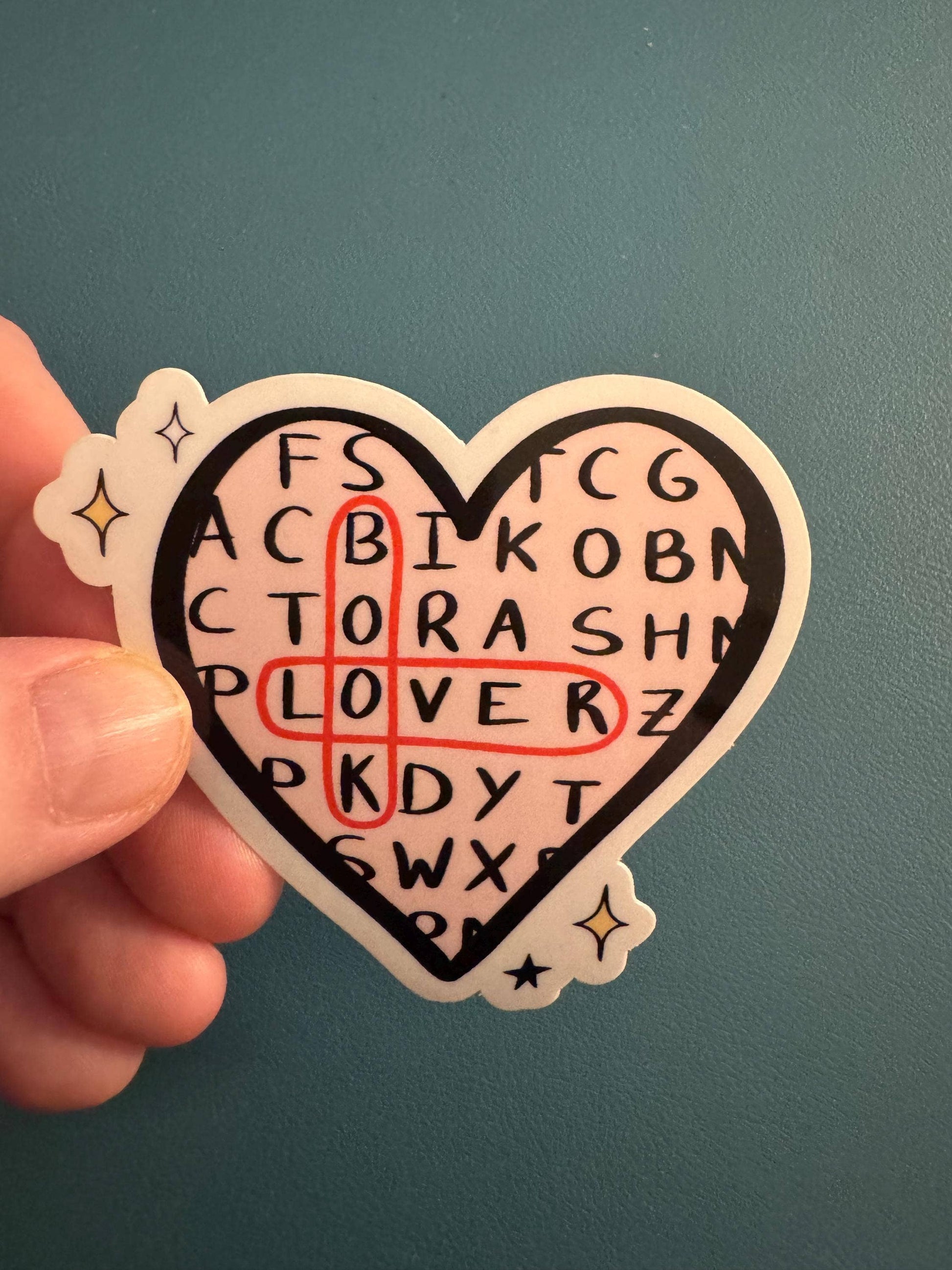 A hand holding a heart-shaped sticker with a crossword puzzle design. The sticker features a black heart outline, a light pink background, black letters spelling out 'LOVER' in the center, and a red crossword intersection drawn over the word.