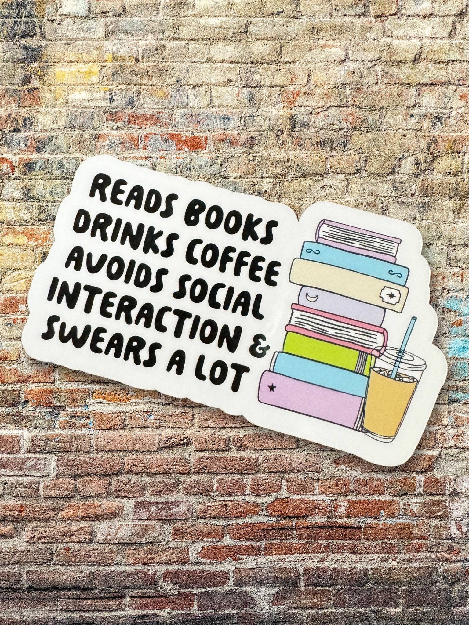Sticker with text 'Reads books, drinks coffee, avoids social interaction & swears a lot' on a brick wall background