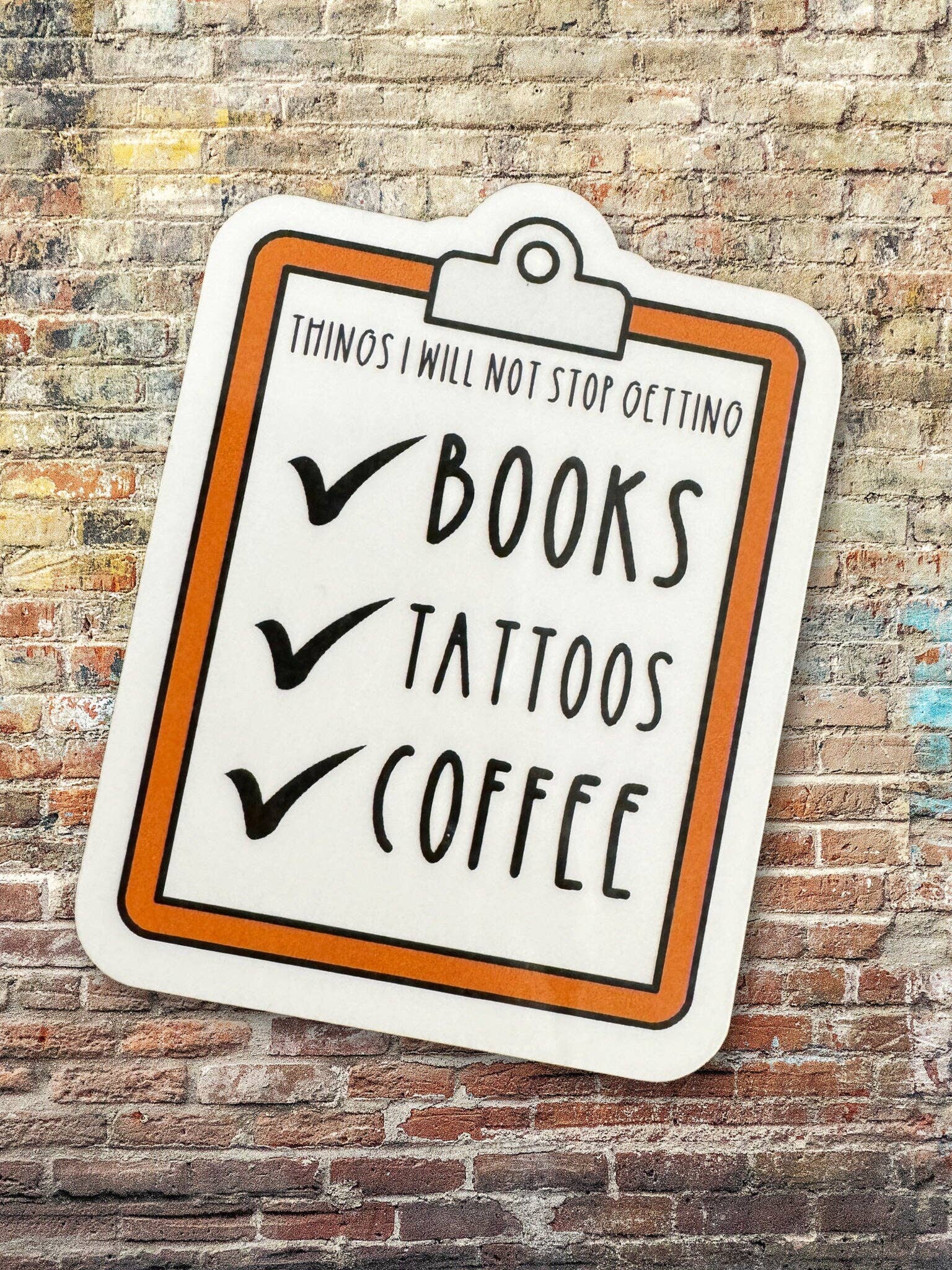 Sticker with 'Books, Tattoos, Coffee' text on a brick wall background