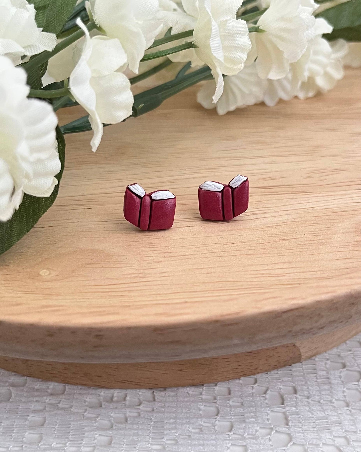 Pair of red book-shaped stud earrings on a wooden surface with white flowers in the background