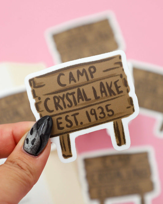 Sticker of 'Camp Crystal Lake' with a hand holding it against a pink background