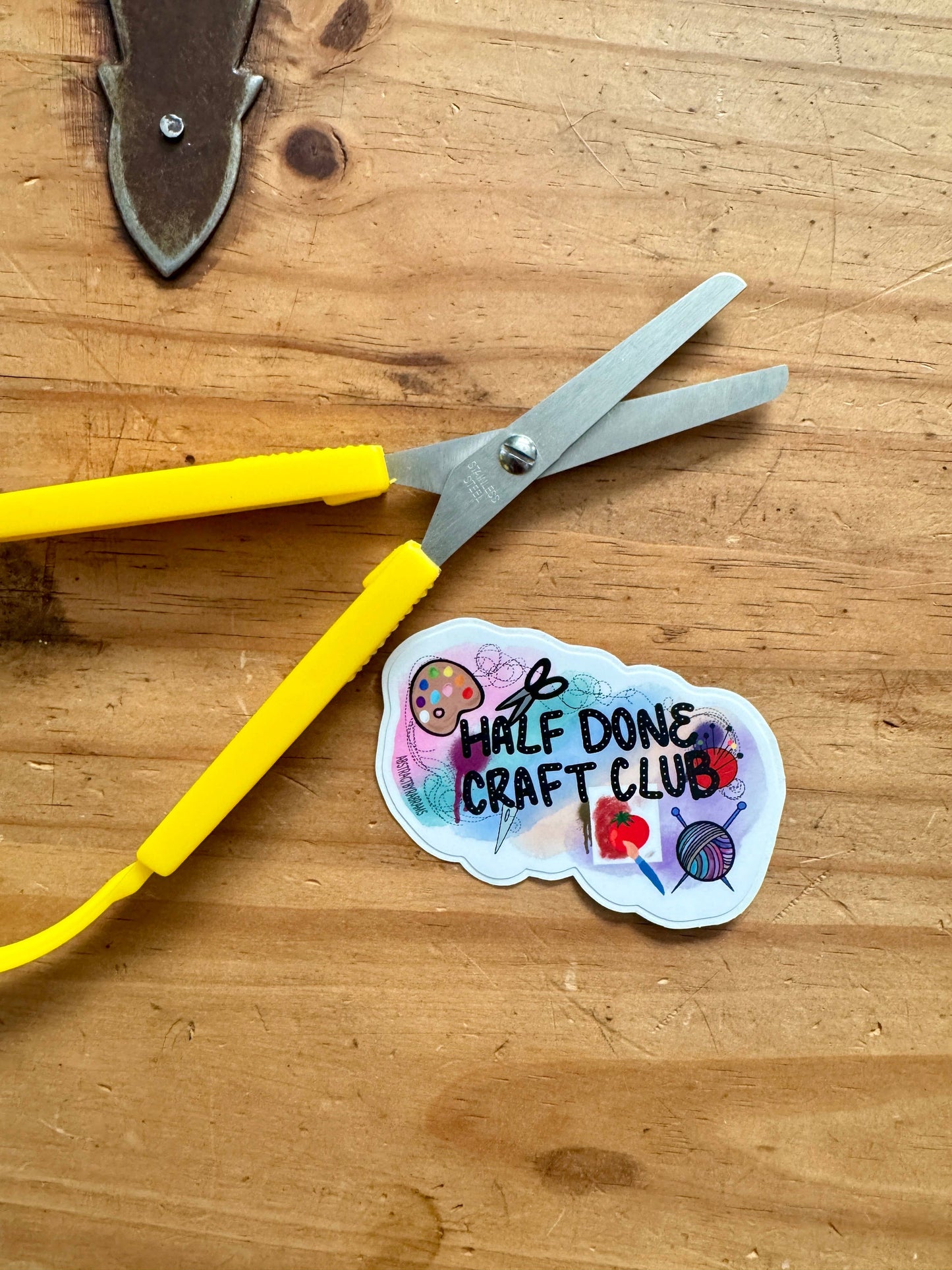 Scissors with a 'Half Done Craft Club' sticker on a wooden surface
