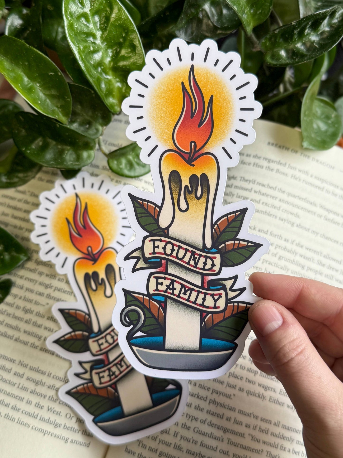 Sticker of a candle with 'found family' text held over a book with green leaves in the background