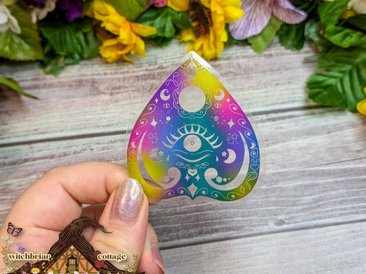 Colorful teardrop-shaped sticker with mystical designs held by a hand on a wooden surface with flowers in the background.