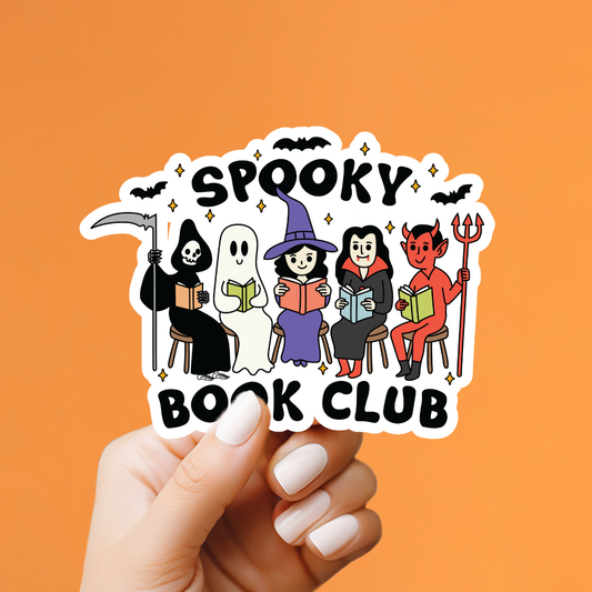A hand with white manicured nails holds a die-cut sticker featuring cartoon characters from a book club: a grim reaper, ghost, witch, vampire, and devil, with the words 'Spooky Book Club' on an orange background.