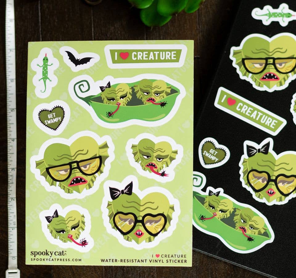 A sheet of cute monster stickers on a light green background, displayed next to a black sheet of stickers on a dark wooden table. The stickers feature a green monster with glasses, bows, and a heart-shaped face, along with text like 'I <3 CREATURE' and 'I <3 SPRAWL'.