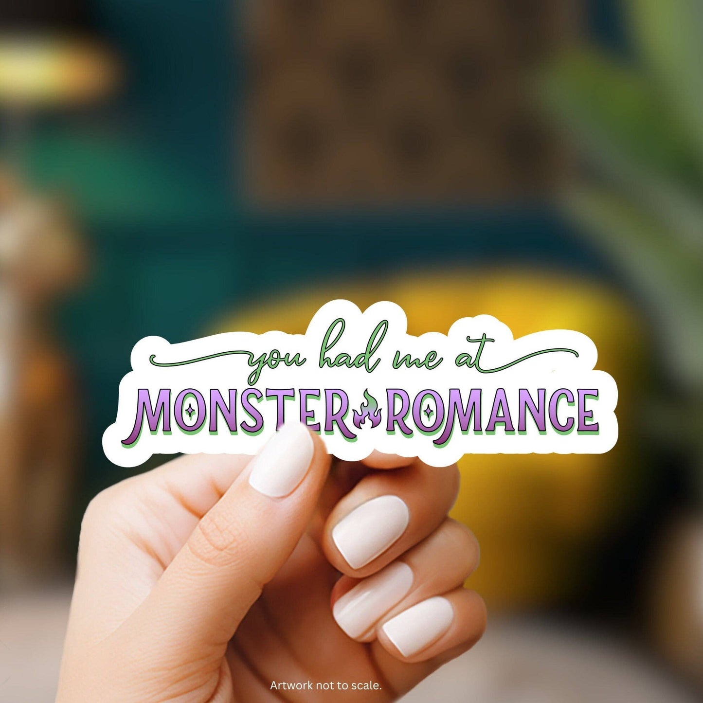 Hand holding a sticker with 'you had me at Monster of Romance' text against a blurred background