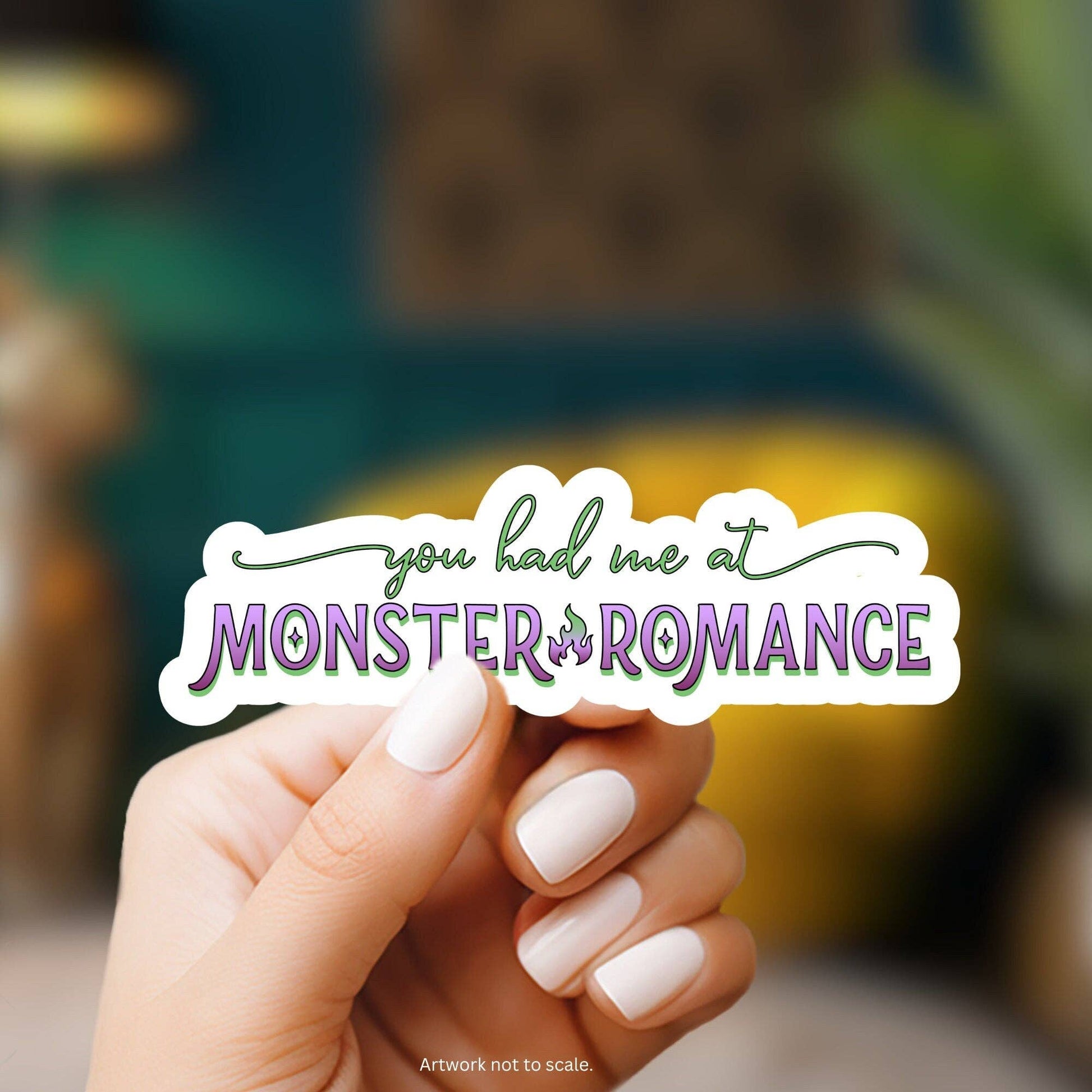 Hand holding a sticker with 'you had me at Monster of Romance' text against a blurred background