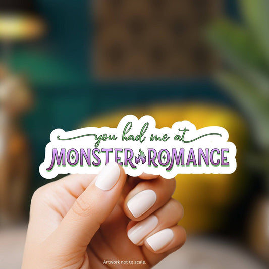 Hand holding a sticker with 'you had me at Monster of Romance' text against a blurred background