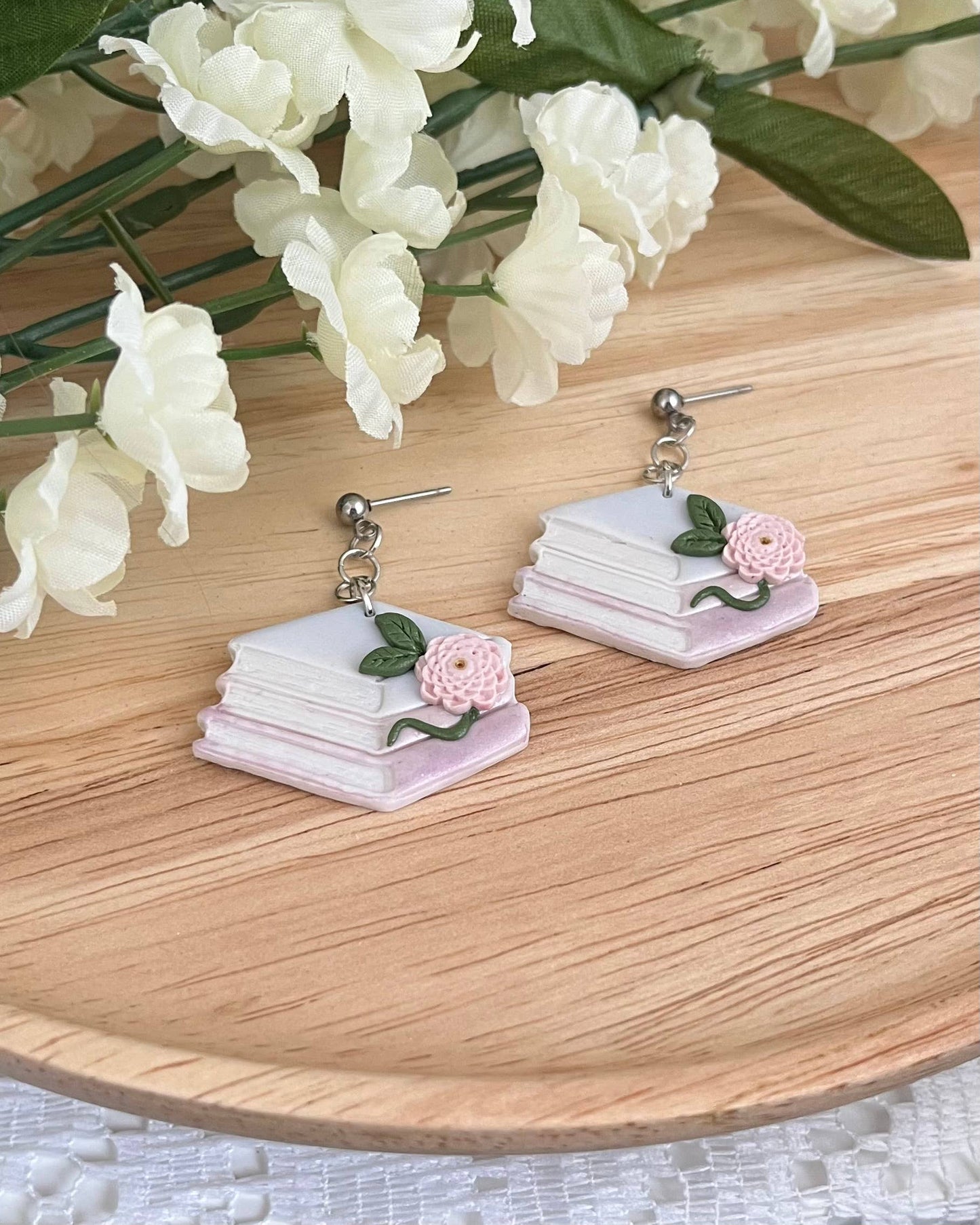 Book-themed earrings with flowers on a wooden surface with white flowers in the background