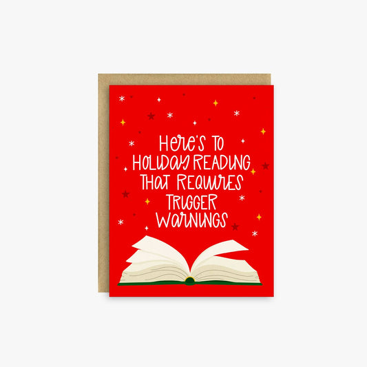 A red greeting card with white hand-lettered text reading 'Here's to Holiday Reading That Requires Trigger Warnings' above an illustration of an open book, shown with a kraft envelope on a light gray background.