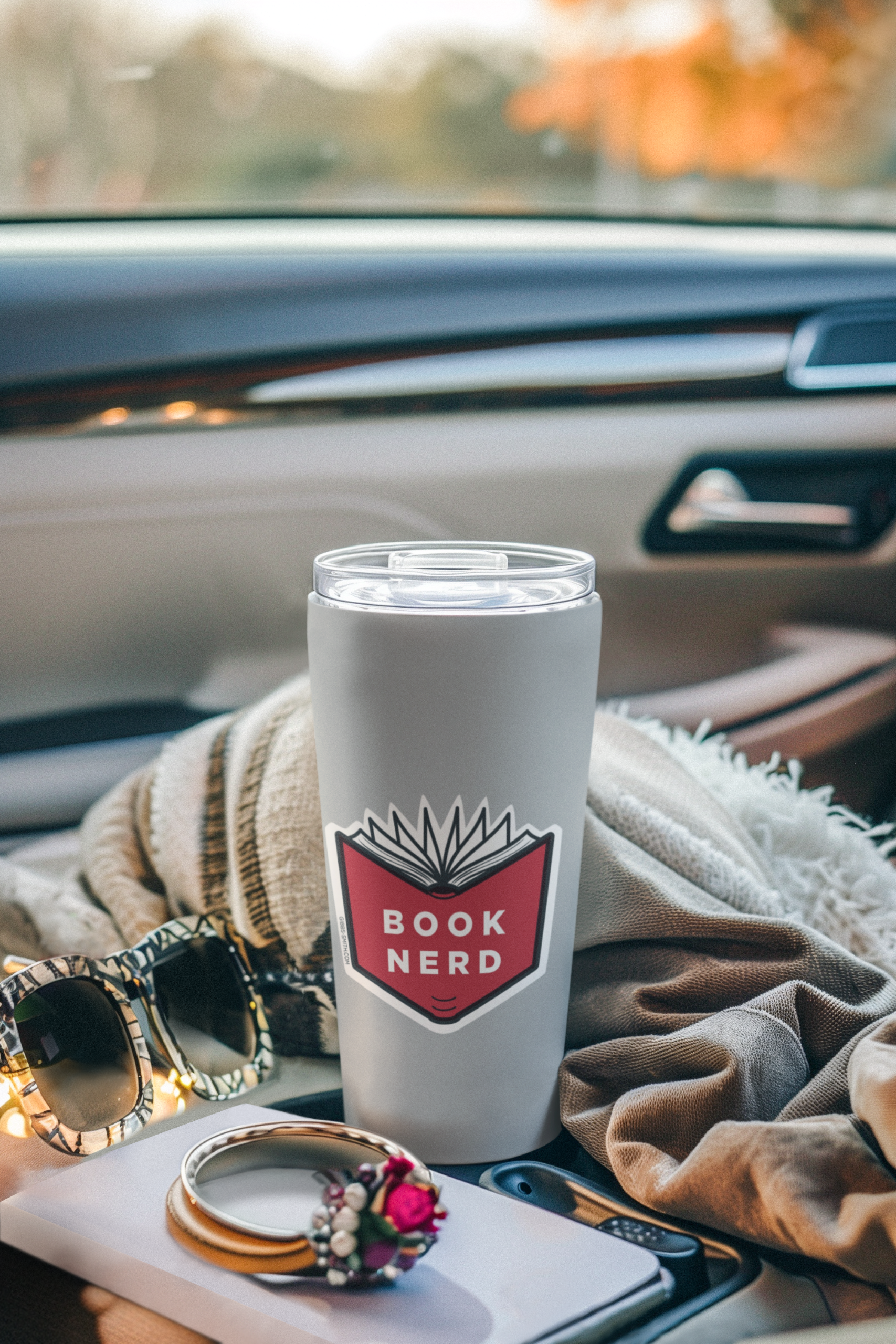 Tumbler with 'Book Nerd' design in a car interior with sunglasses and notebook.