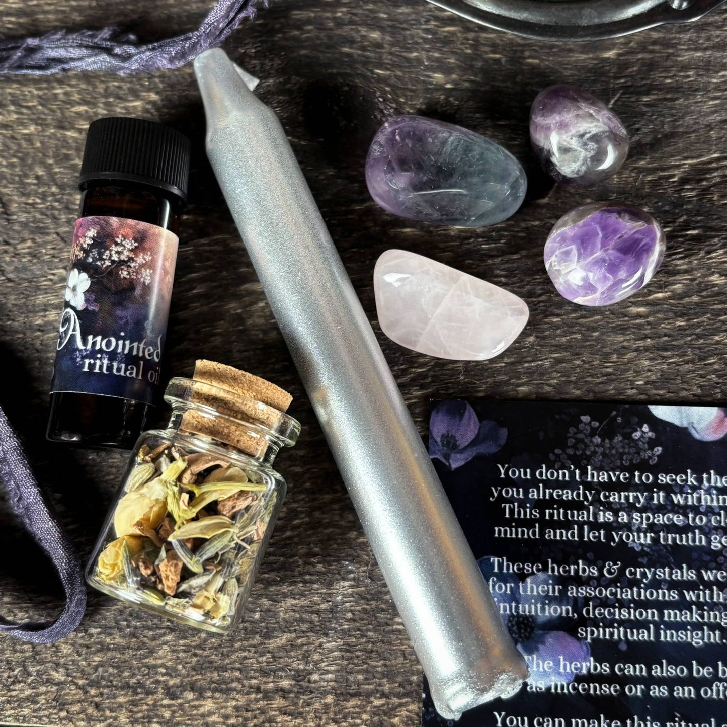 Herbal concoction in a small jar, bottle labeled 'Anointed Ritual Oil', and crystals on a wooden surface.