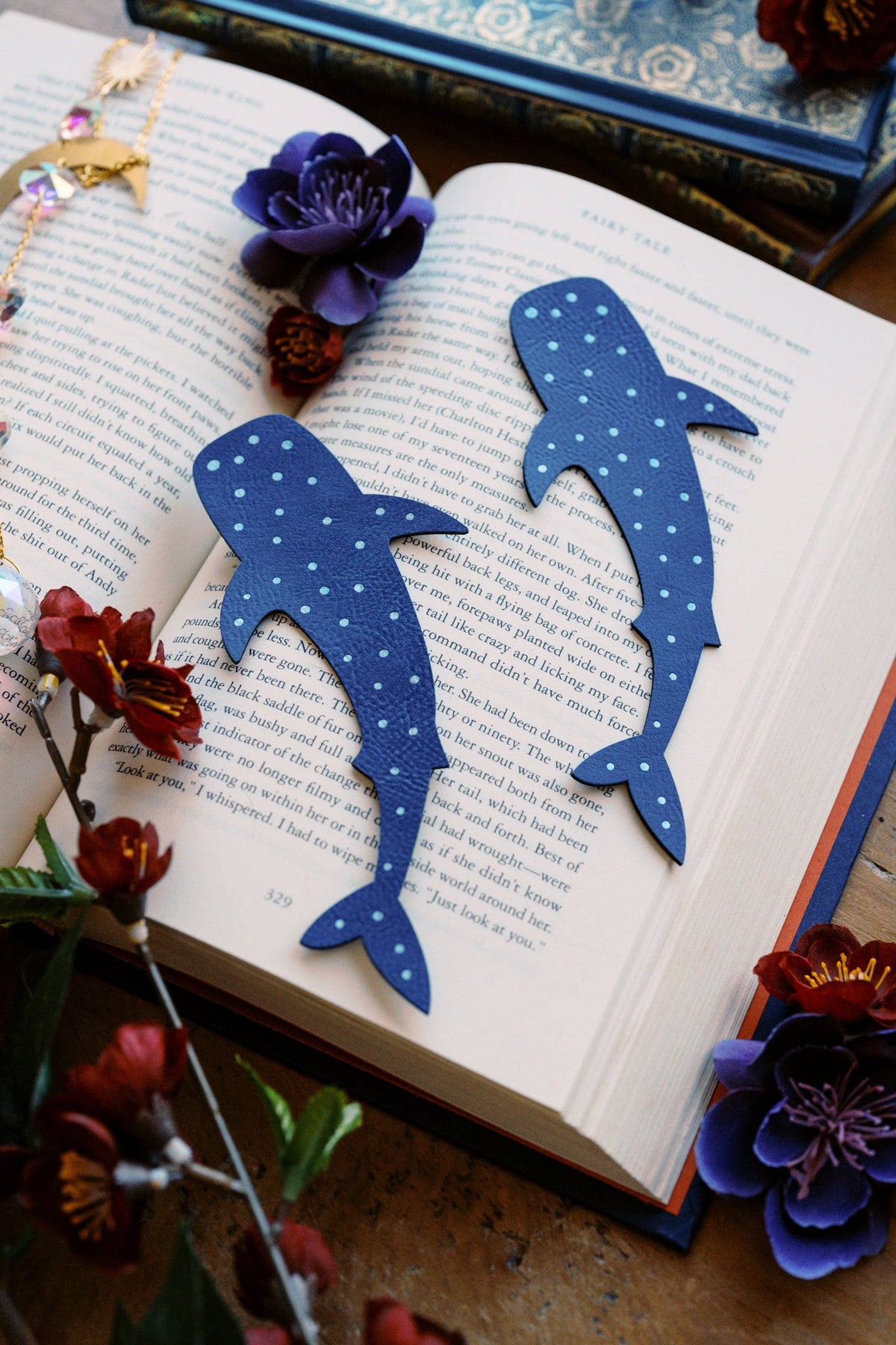 Two blue shark-shaped bookmarks with white polka dots resting on an open book, surrounded by decorative flowers and books.