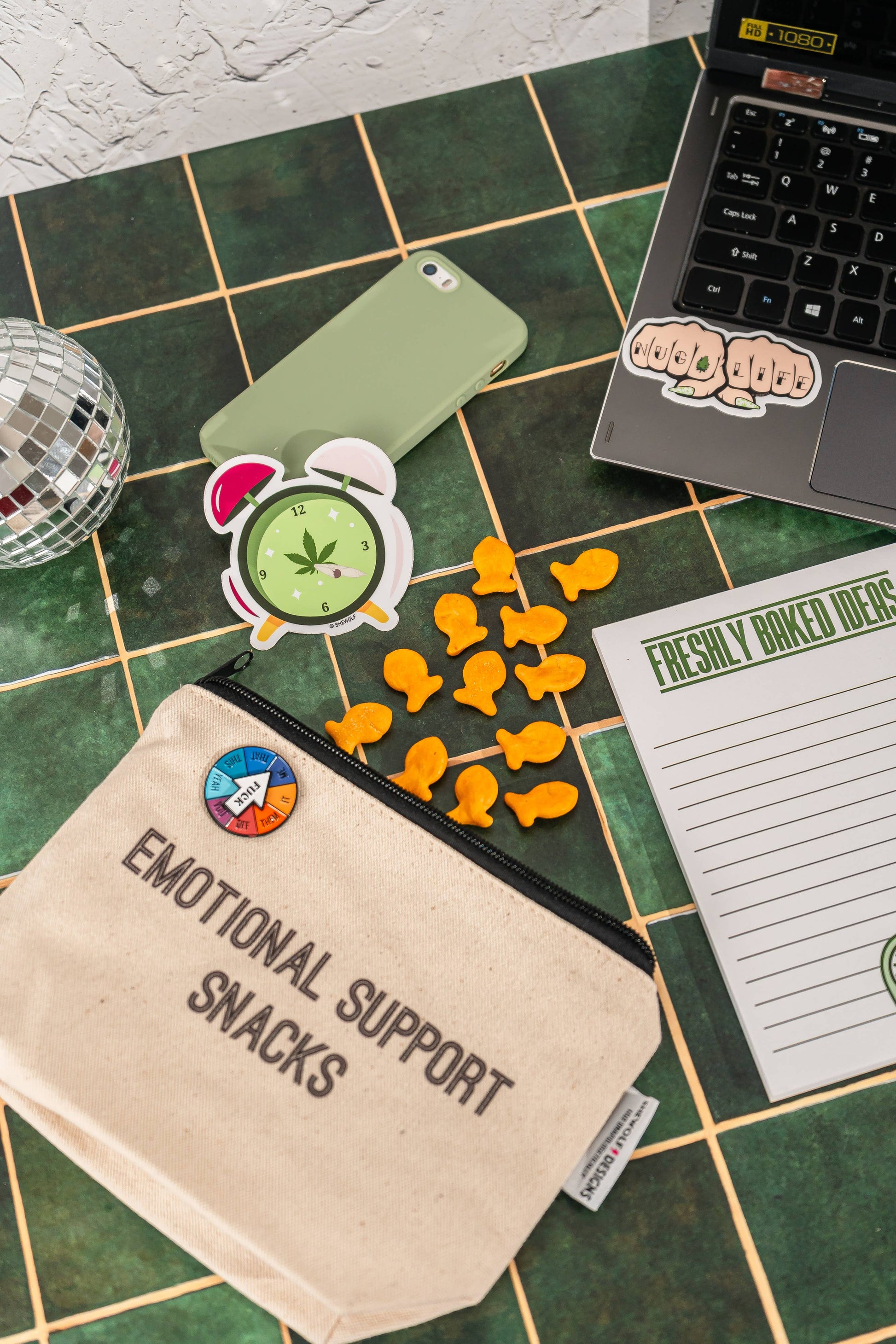 Desk with 'Emotional Support Snacks' bag, phone, laptop, and notepad on a green tiled surface.