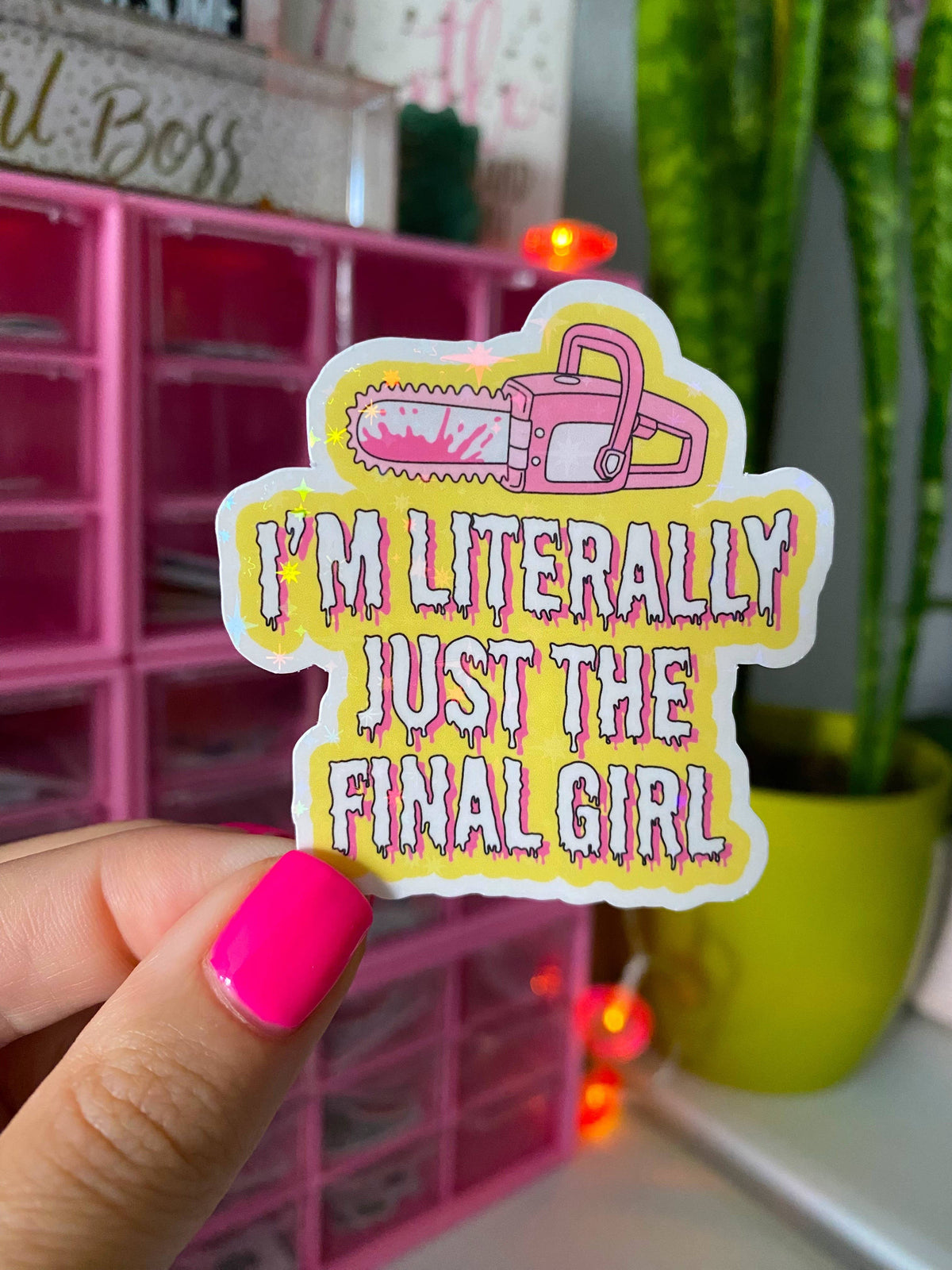 A hand with bright pink nail polish holds a die-cut sticker featuring a pink chainsaw and the text 'I'M LITERALLY JUST THE FINAL GIRL' in dripping pink letters on a yellow background. The scene is set in a room with a pink organizer and a green plant in the background.