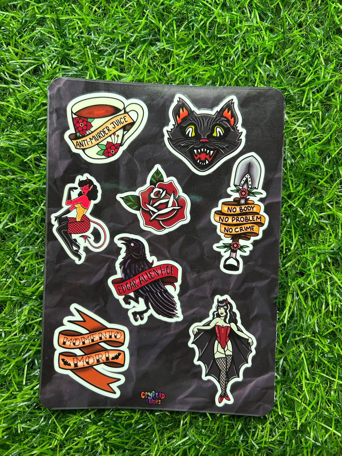 A sheet of nine colorful tattoo-style stickers displayed on a black backing, laid flat on green grass. The stickers feature a black cat, a red rose, a raven, a woman in a red dress, a teacup with a cat inside, and banners with text including 'ENTROPY WICK' and 'NO PROBLEM NO CARE'.
