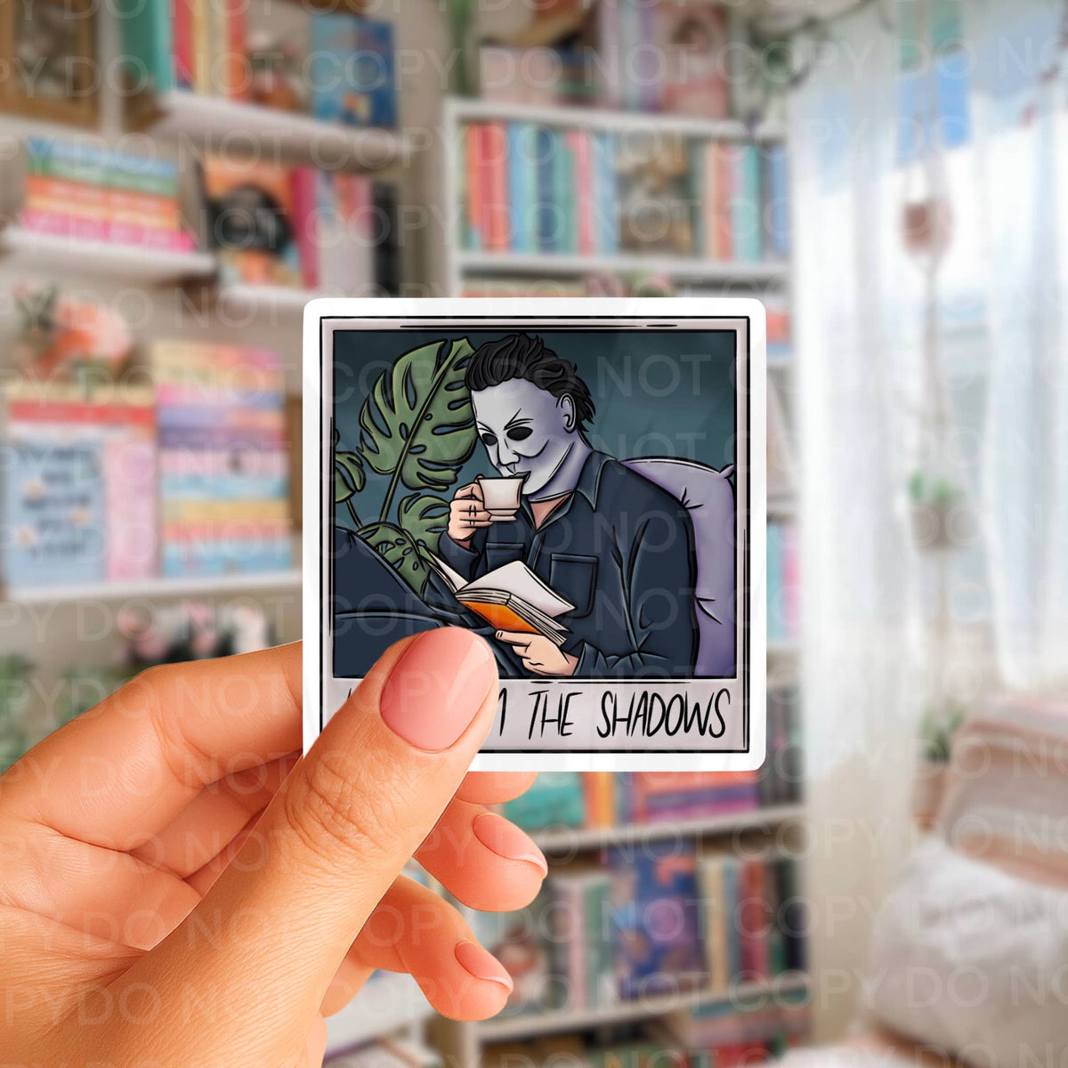 A hand holds a square sticker featuring an illustrated character in a mask reading under a plant, with the text 'I THE SHADOWS' at the bottom. The background is a blurred bookshelf scene.