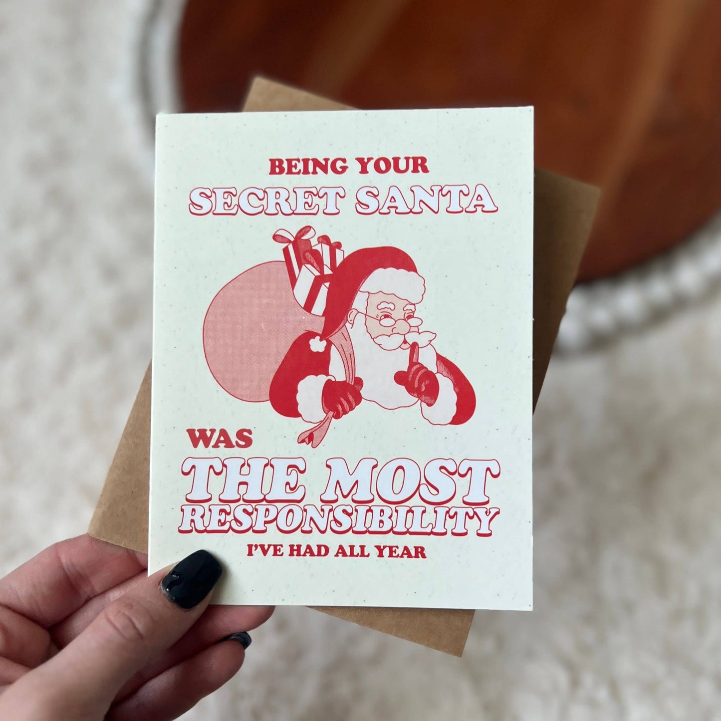 A hand with black nail polish holds a white greeting card featuring a red illustration of Santa carrying gifts, with the text 'BEING YOUR SECRET SANTA WAS THE MOST RESPONSIBILITY I'VE HAD ALL YEAR'. A brown envelope is partially visible behind the card, against a soft beige background.