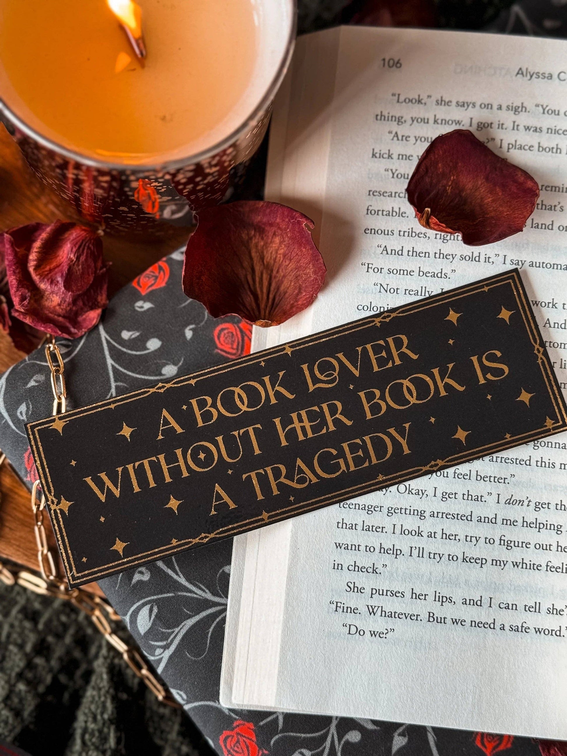 Book with a bookmark reading 'A book lover without her book is a tragedy', surrounded by dried roses and a lit candle.