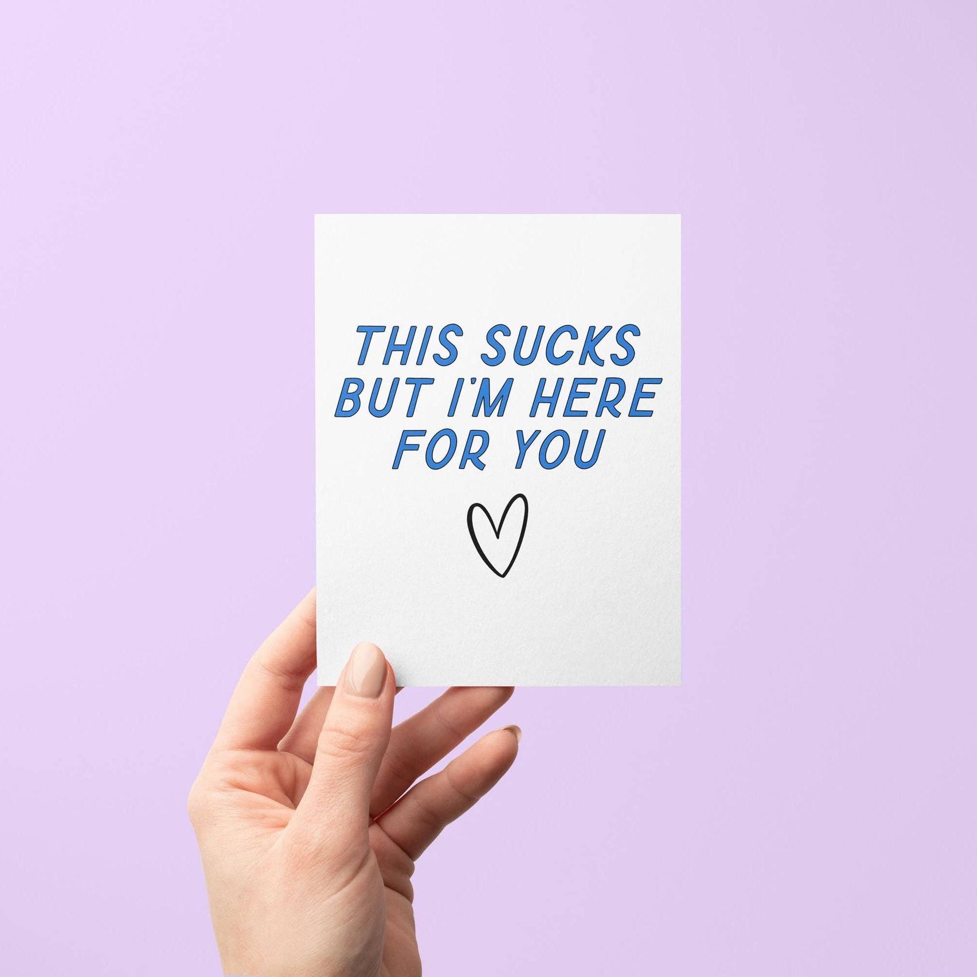 Hand holding a card with 'This sucks but I'm here for you' text on a purple background