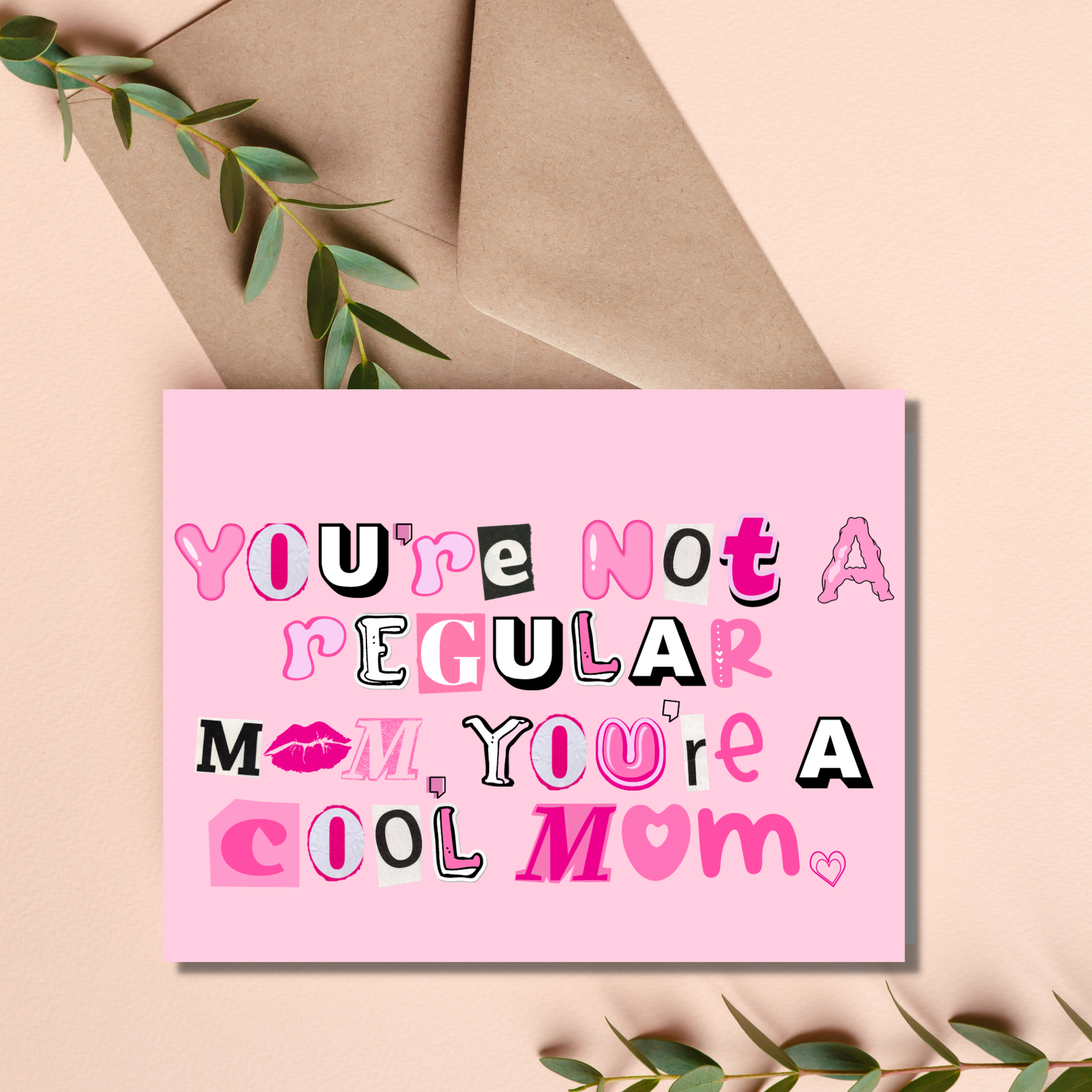 Pink card with text 'You're not a regular mom, you're a cool mom' on a beige background with an envelope and green leaves.