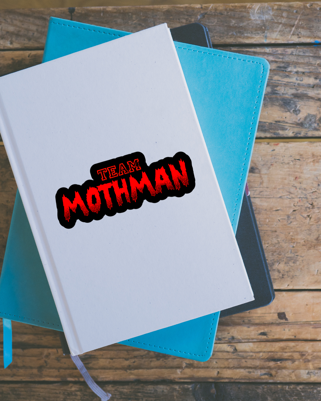Notebook with 'Team Mothman' sticker on a wooden surface