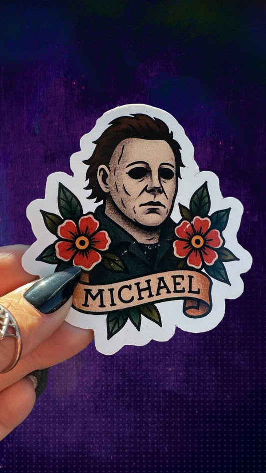 A hand holds a die-cut sticker featuring a portrait of Michael Myers with red flowers and the name 'MICHAEL' on a banner, against a dark purple textured background.
