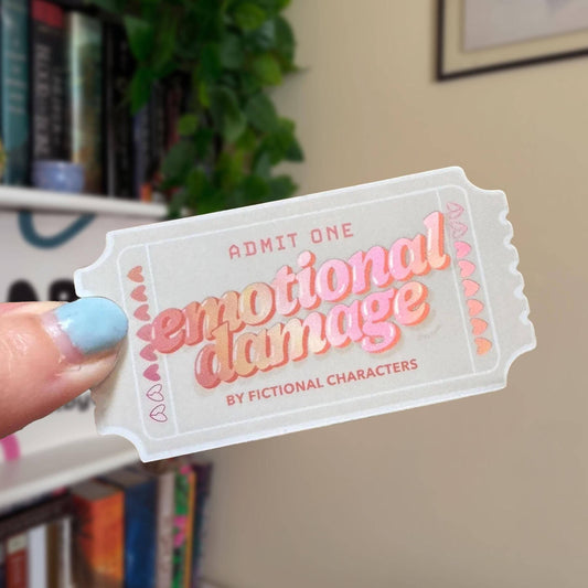Ticket-shaped sticker with 'emotional damage' text held by a hand against a blurred bookshelf background