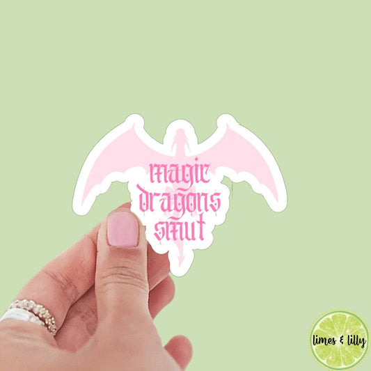 Pink dragon-shaped sticker with 'magic dragons slay' text held by a hand against a light green background.