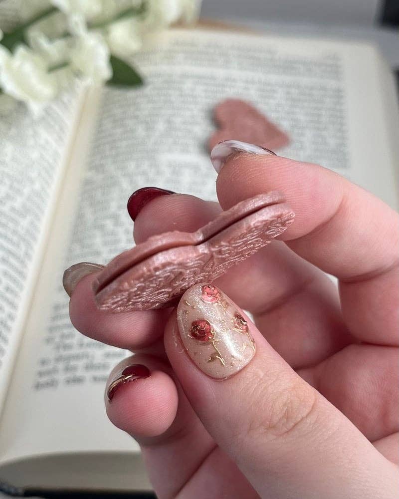 Hand holding a rose gold corner bookmark with a blurred background of an open book and flowers.