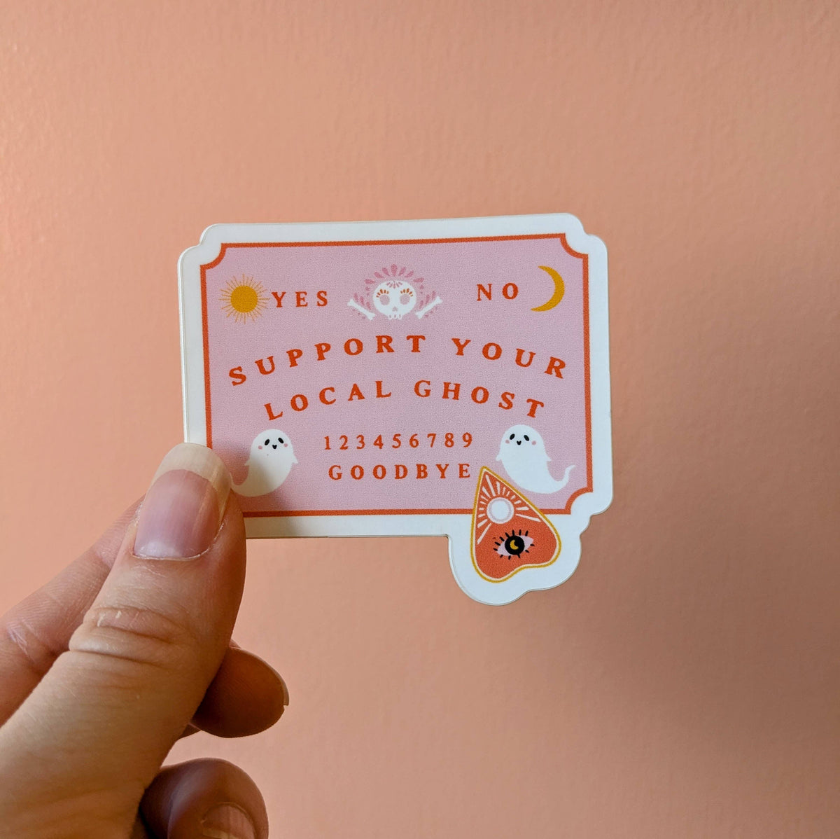 A hand holds a die-cut sticker featuring a pink and orange gothic-style ghostly Ouija board design with the text 'SUPPORT YOUR LOCAL GHOST' and small white ghost illustrations, against a solid peach-pink background.