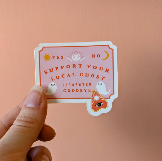 A hand holds a die-cut sticker featuring a pink and orange gothic-style ghostly Ouija board design with the text 'SUPPORT YOUR LOCAL GHOST' and small white ghost illustrations, against a solid peach-pink background.