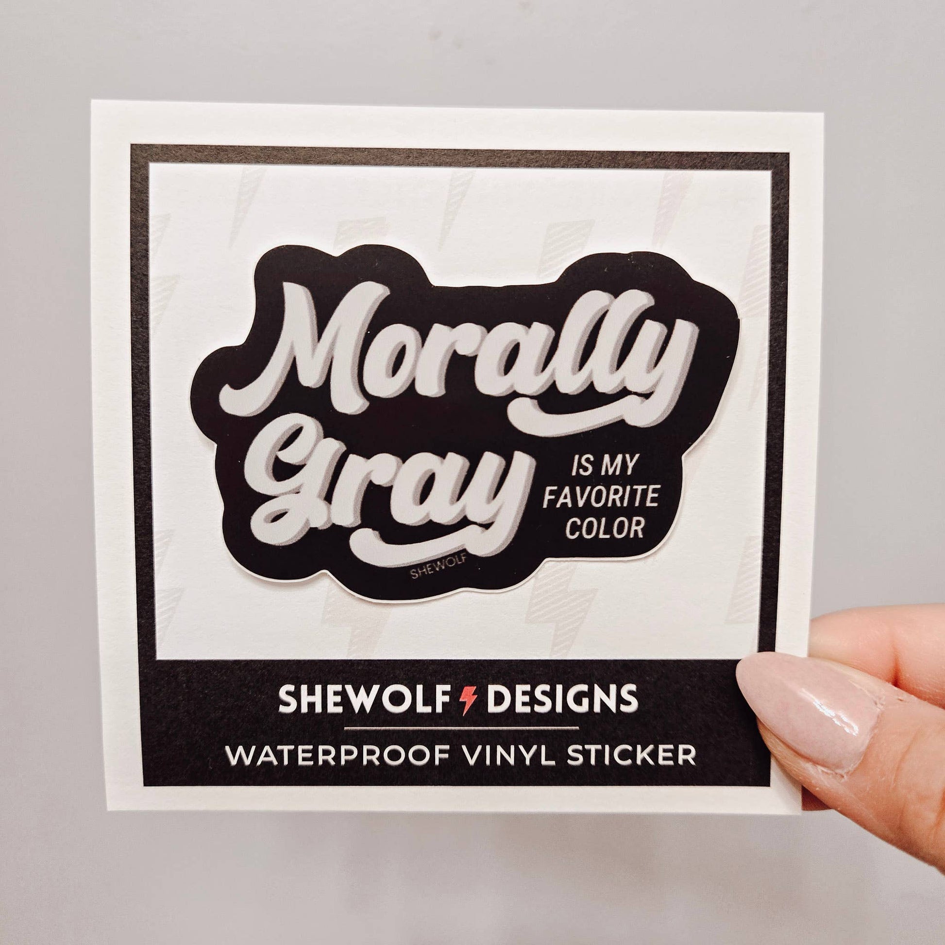 Sticker with 'Morally Gray is my favorite color' text held by a hand on a gray background