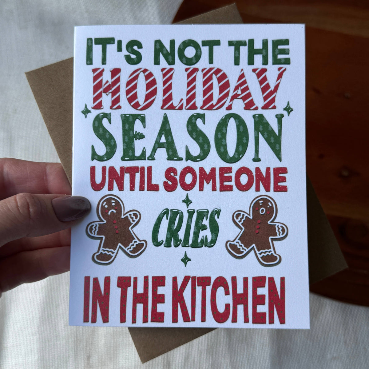 A hand holds a white Christmas greeting card with the text 'IT'S NOT THE HOLIDAY SEASON UNTIL SOMEONE CRIES IN THE KITCHEN' in green and red lettering, alongside two gingerbread cookies. A brown envelope is visible behind the card.