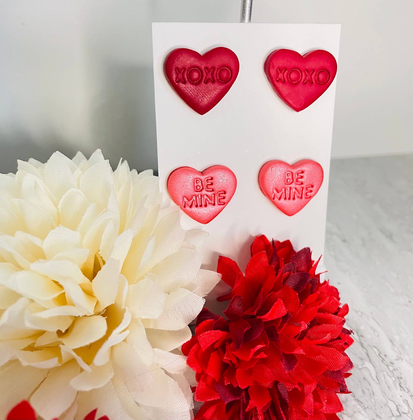Heart-shaped earrings with 'XOXO' and 'Be Mine' text on a card, surrounded by flowers.