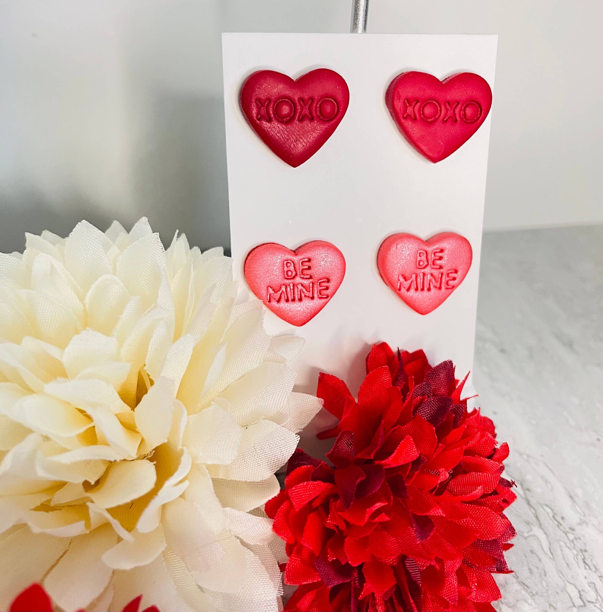 Heart-shaped earrings with 'XOXO' and 'Be Mine' text on a card, surrounded by flowers.