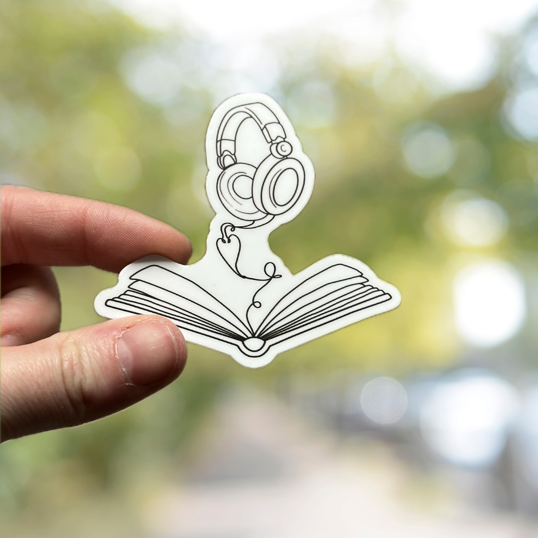 A hand holds a die-cut sticker featuring a black line art illustration of over-ear headphones above an open book, set against a blurred green outdoor background.
