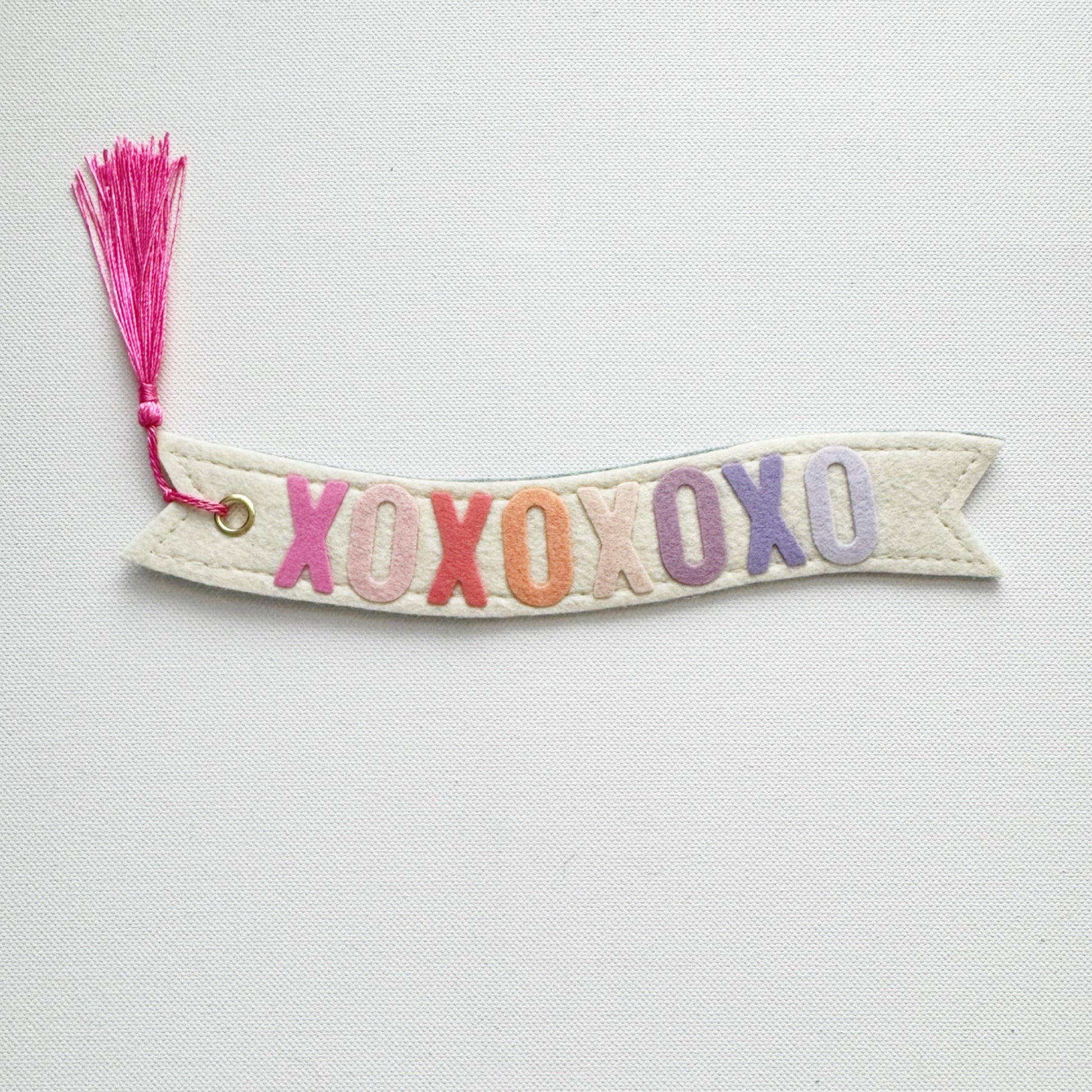 Banner-shaped bookmark with 'XOXO' in colorful letters and a pink tassel on a light gray background