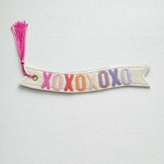 Banner-shaped bookmark with 'XOXO' in colorful letters and a pink tassel on a light gray background